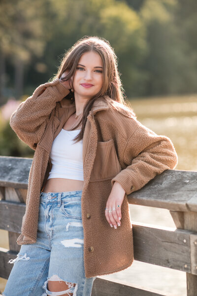 Sun-filled image of high school senior girl in off-white off the shoulder sweater leaning on wooden fence