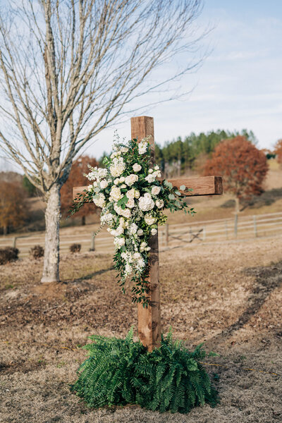 wooden cross standing outside with a bush of greenery at the bottom and florals in the center of it