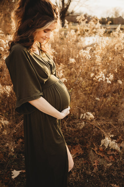 A mother stands in a warm grass field smiling down at her pregnancy bump.