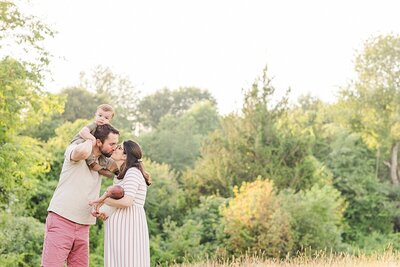 boy sits on dad shoulders while parents kiss  during outdoor newborn photo session with Sara Sniderman Photography at Oak Grove Park in Millis Massachusetts