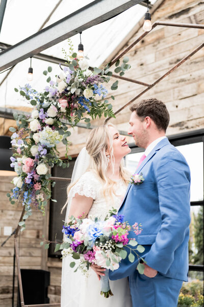 Bride and groom looking and smiling at each other under a flower arch and holding a matching flower bouquet