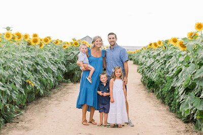 a mom wearing a blue dress holding a toddler boy  standing next to a dad in a blue shirt and a little girl in a white dress and a little boy in a blue shirt stand in the middle in a sunflower field by seattle wa family photographers Shaunae Teske