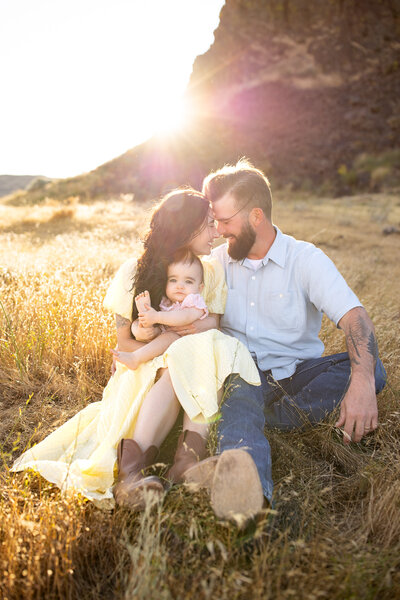 Parents snuggling close with their baby in a sunlit field, sharing a quiet, joyful moment together.