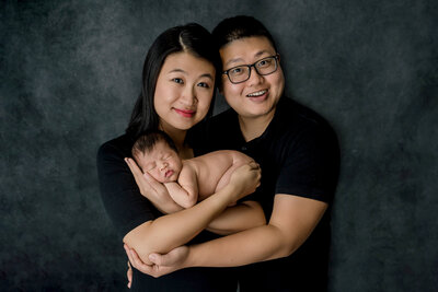 Joyful new parents smiling while holding their sleeping newborn baby wrapped in white during professional studio portrait session on dark backdrop