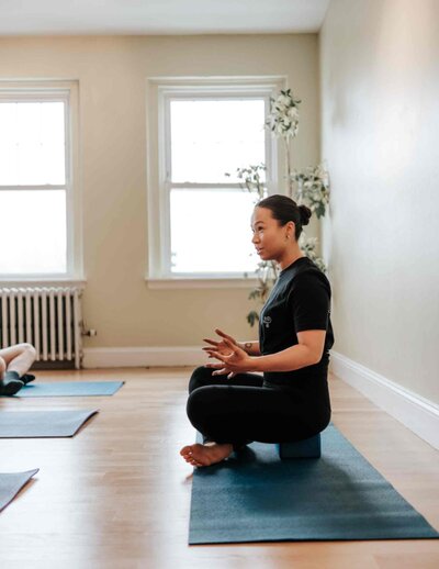 A breathwork instructor sits cross legged in front of a group of participants, explaining how to do deep breathing techniques in a sunlit studio.