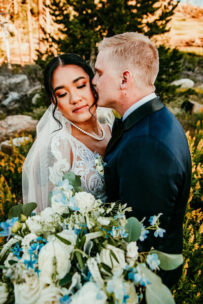 A groom gently kisses his bride's cheek in a tender, intimate moment amidst a meadow of yellow wildflowers, with lush greenery surrounding them in Lake Tahoe