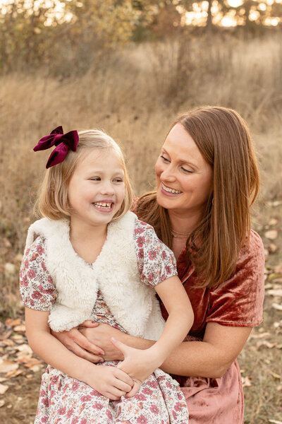 7 year old girl sitting on moms lap outdoors at Portland Family Photo session. Mom is wearing a velvet mauve dress and little girl is wearing a mauve floral dress with white fuzzy vest and a big mauve bow in her hair. Outdoors in a tall golden grass field.
