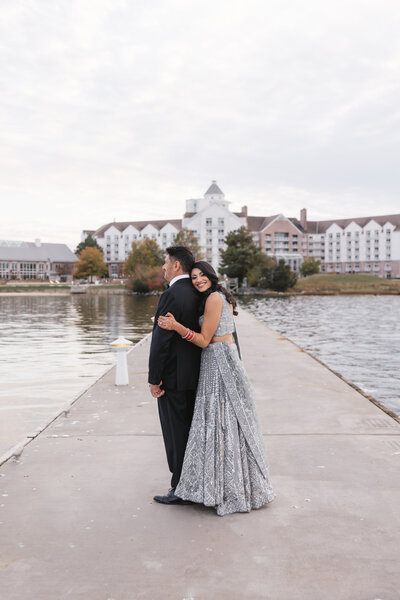 Bride in silver lehenga hugging groom during waterfront South Asian wedding portraits on a dock at sunset.