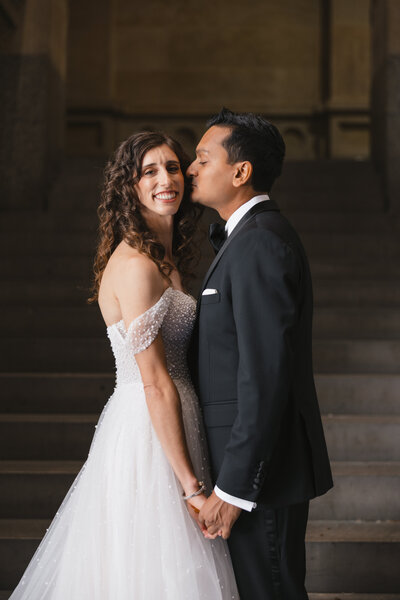 Sweet couple in wedding attire holds hands while the groom gives the bride a kiss.