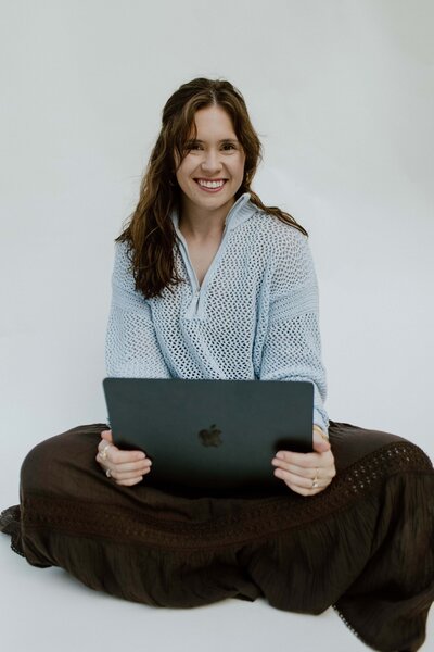 A woman sitting in front of a white backdrop with her laptop on her lap smiling.