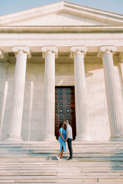A couple kissing in front of a building with tall stone pillars 