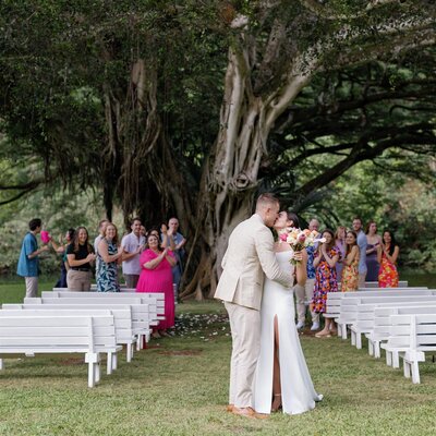 A newlywed couple kissing in the middle of the aisle while their wedding guests cheer behind them 