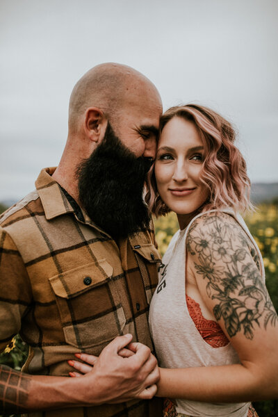 husband and wife couple nuzzled in for a sweet moment smiling in yellow daisy flower field with beard and tattoos