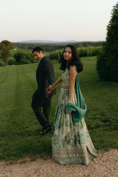An Indian couple, the man is wearing a navy blue suit, and the woman is wearing a green lengha. They are holding hands walking through the winery with greenery background. The woman is looking back at the camera.