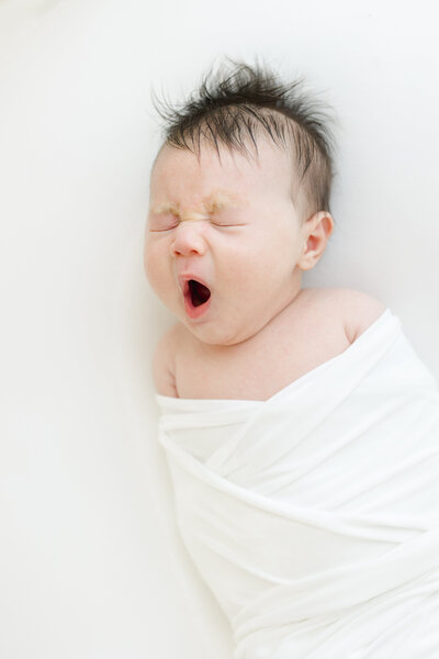Swaddled newborn baby yawns during newborn photography session