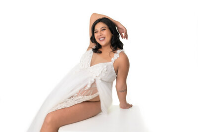 Maternity picture of a pregnant mom sitting on a posing box with her hand above her head, smiling.