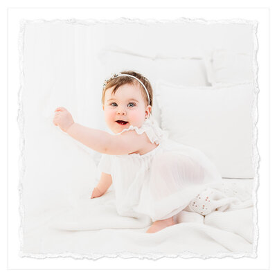 A captivating photograph captures the essence of a joyful moment. A one-year-old girl smiles brightly, radiating happiness. Against a pure white background, she stands confidently, radiating purity and grace. This milestone photo session by Bri Sullivan Photography beautifully captures the essence of childhood innocence.