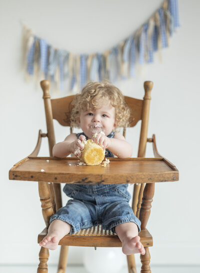 Toddler with curly hair enjoying a cake smash in a vintage wooden high chair during a milestone session with a Kansas City family photographer.