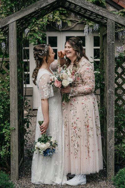 An LGBTQ+ couple candidly pose together for wedding photography in a garden. They both wear dresses and smile.