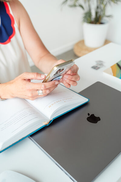 Calgary brand storytelling photoshoot of a woman using her smartphone at a clean, modern workspace. Her hands rest on an open planner with ‘Weekly Preview’ printed on the page, next to a sleek MacBook. A few SD memory cards and a notebook are scattered on the white desk, with a potted plant in the softly blurred background, creating a productive and organized atmosphere.