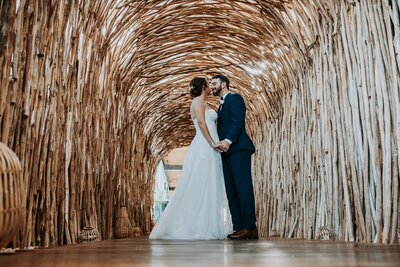 A bride and groom share a kiss while standing under a woven wooden archway. The bride wears a lace wedding gown, and the groom is dressed in a navy blue suit as they hold hands.