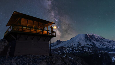 A woman stands on Fremont Lookout at Mt Rainier with a lantern under the Milky Way