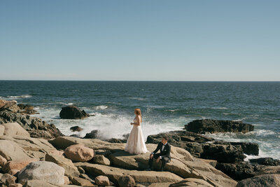 A bride standing looking out at the ocean and a groom sitting looking at the bride overlooking waves at Acadia National Park in Maine. 