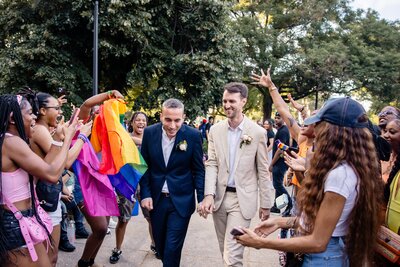 Two husbands walk through a park while a pride flag blows in the air during a pride parade
