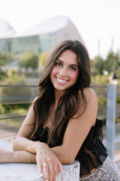 A woman in a white dress and sash holds a graduation cap outdoors. She's smiling, with sunlight filtering through trees, creating a joyful tone.