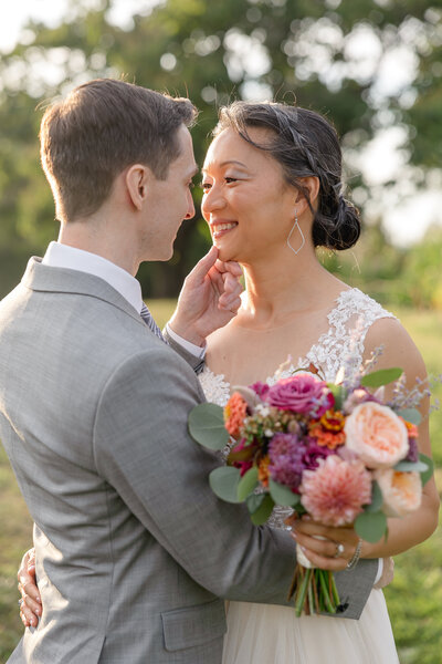 A bride wearing a white lace dress embraces her husband in a grey suit and white shirt as she holds onto a bouquet of bright flowers while smiling