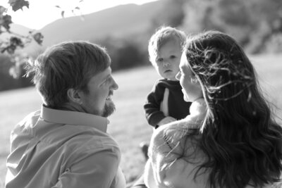A black-and-white photo of three people in a park captures the essence of a New England Family Photographer. An adult with short hair smiles at another adult with long hair, who is holding a child. The child looks towards the camera as sunlight filters through trees, creating a serene atmosphere.