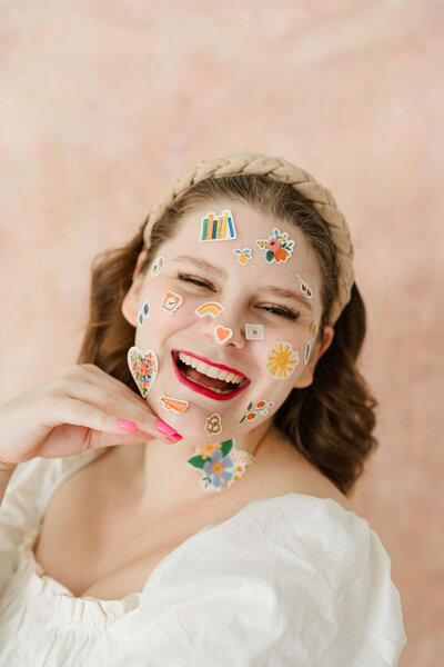 headshot of copywriter wearing red lipstick, with stickers all over her face, and pink nail polish, smiling at the camera