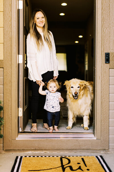 mom holds hand of baby girl by front door as blonde dog stands next to them
