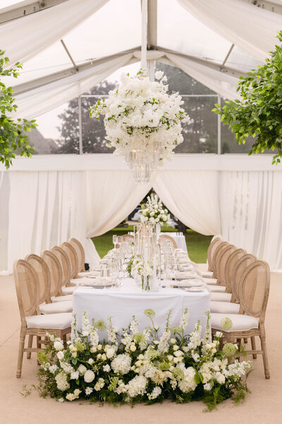 Wedding reception in a text, long rectangle tabled with a white table cloth, wood cushioned chairs, white bouquets hanging above , and at the end of the table there are white flowers and greenery on the floor