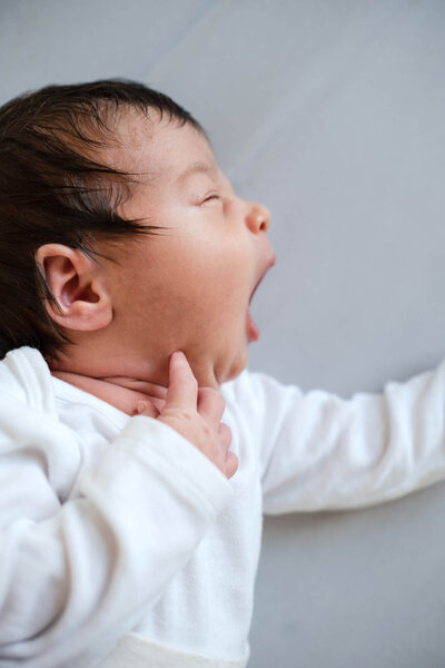 A sleepy baby in white clothing yawns widely while lying down on a light background.