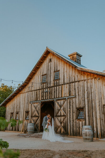 bride and groom kiss in front of rustic barn wedding venue in northern california