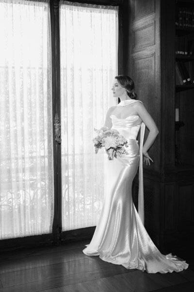 Aldrich Mansion Wedding Photos | Kelsey Sheehan Photography Timeless Rhode Island Weddings | A woman in a sleek, satin bridal gown stands elegantly by a large window, holding a bouquet. The black-and-white photo conveys a timeless, serene mood.