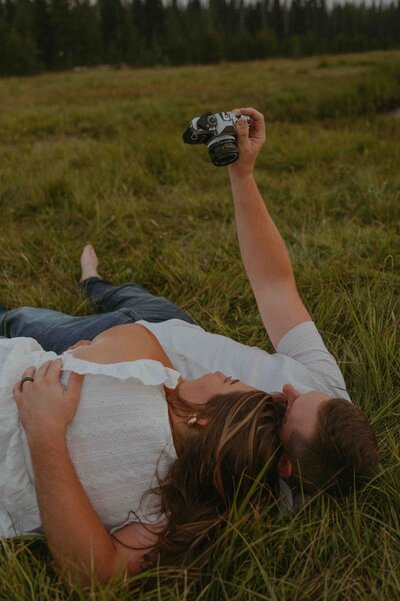 Couple laying in a field, her head on his shoulder while holding a film camera above them for a photo