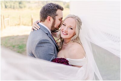 close up of groom leaning into brides temple while she looks at the camera holding her deep red bouquet and the veil wraps around her covering the foreground