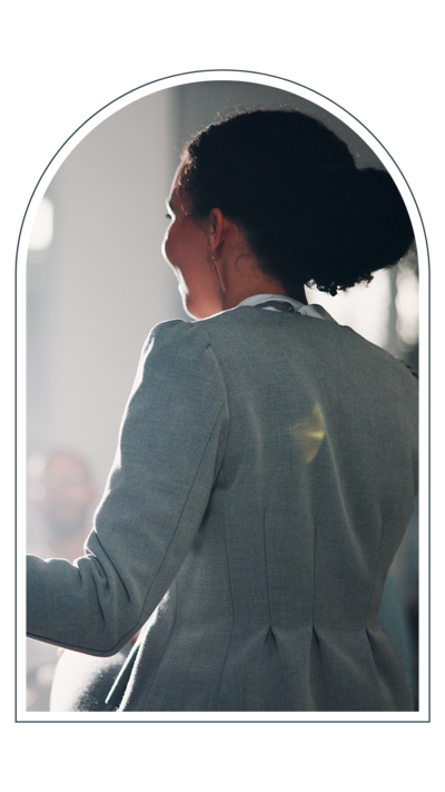  A woman in a gray blazer, seen from behind, speaking to an audience in a bright room, with sunlight highlighting her silhouette.