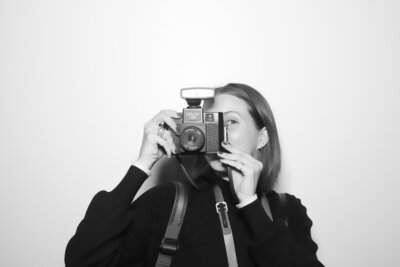 Black and white image of Bailee holding a Holga camera up in front of her face in front of a white wall