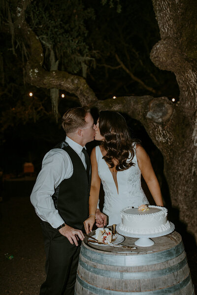 bride and groom kiss while cutting wedding cake at outdoor wedding reception