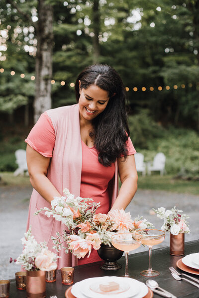 Jasmin in a pink dress making adjustments to a centerpiece  at an outdoor wedding reception in upstate New York
