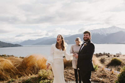 The Lovers Elopement Co - Groom at wedding ceremony ontop of a mountain in Queenstown New Zealand with lake below