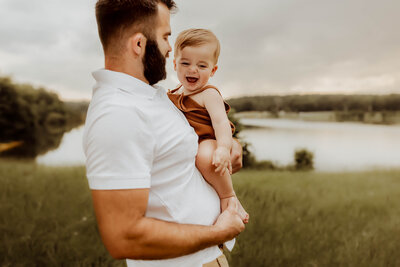 Macon Family Photographer captures a heartwarming moment of a man holding a baby in front of a serene lake.