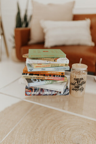 This image shows a stack of books on a glass desk, with an iced coffee next to them in a tumbler that says "Fueled by iced coffee and anxiety."