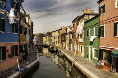 Brightly painted houses line a narrow canal in Burano, Italy, with small boats reflecting in the water.