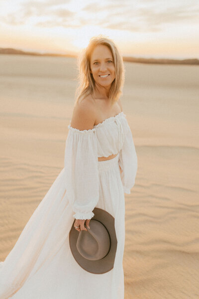 Blonde woman  looking right at the camera, she is wearing a long white skirt with a an off the shoulder matching top. She is on the beach at sunset.