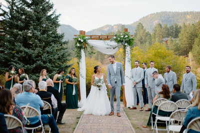 A newlywed bride and groom walk hand-in-hand down the aisle at Mountain View Ranch in Pine, Colorado, celebrating their love and the beginning of their new life together. The rustic charm of the venue adds to the heartfelt moment.