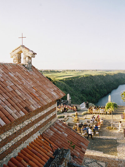 Aerial view of a wedding ceremony at Altos de Chavón, with the stone chapel and Chavón River framed in warm sunlight — destination wedding by Asia Pimentel.